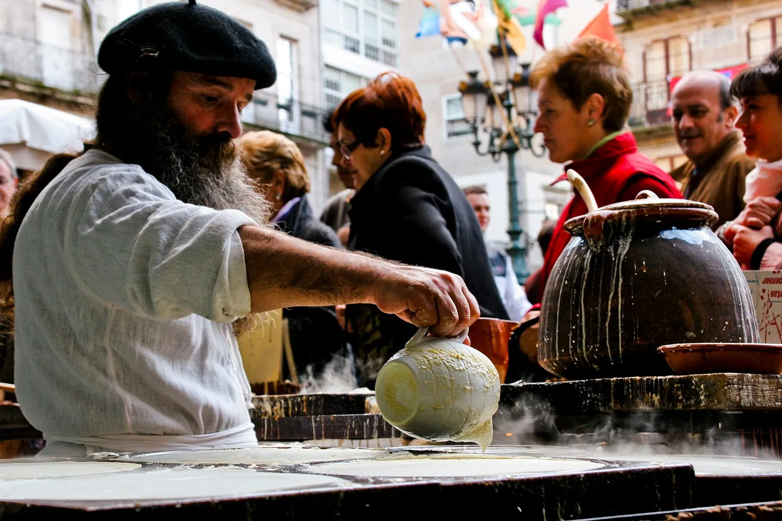Un cocinero vierte masa de filloas sobre una plancha ante un puesto del mercado, con público alrededor