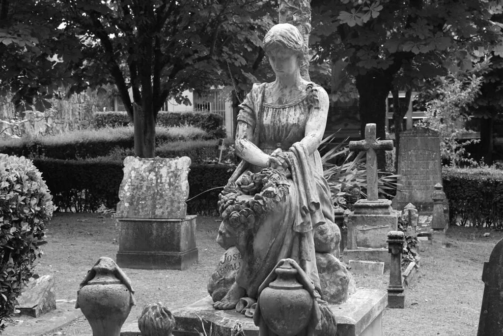 Escultura de una mujer sentada con una corona de flores entre las manos, junto a cruces y urnas