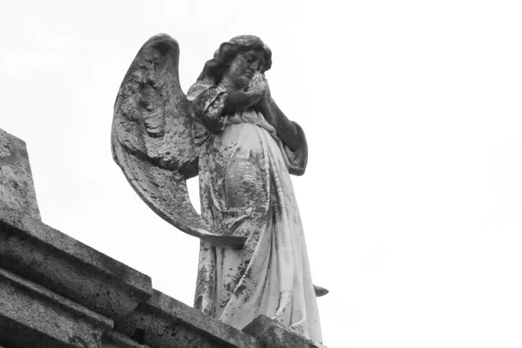 Ángel de piedra con las alas plegadas y la cabeza inclinada sobre un panteón, fotografiado en blanco y negro