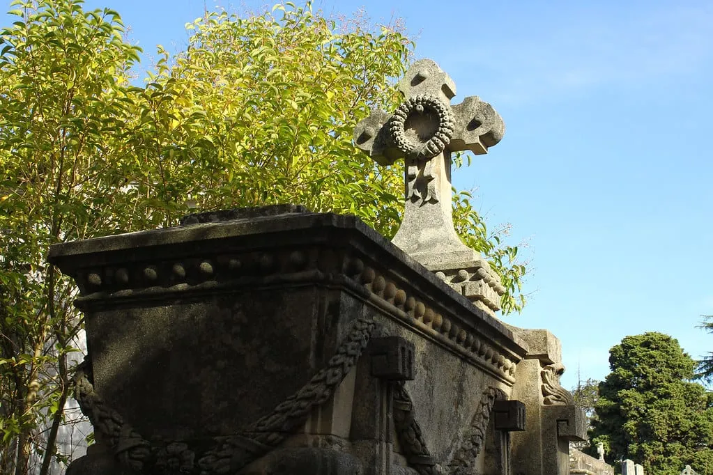 Panteón de piedra con una cruz coronada por una corona de laurel, con árboles de hojas amarillas al fondo