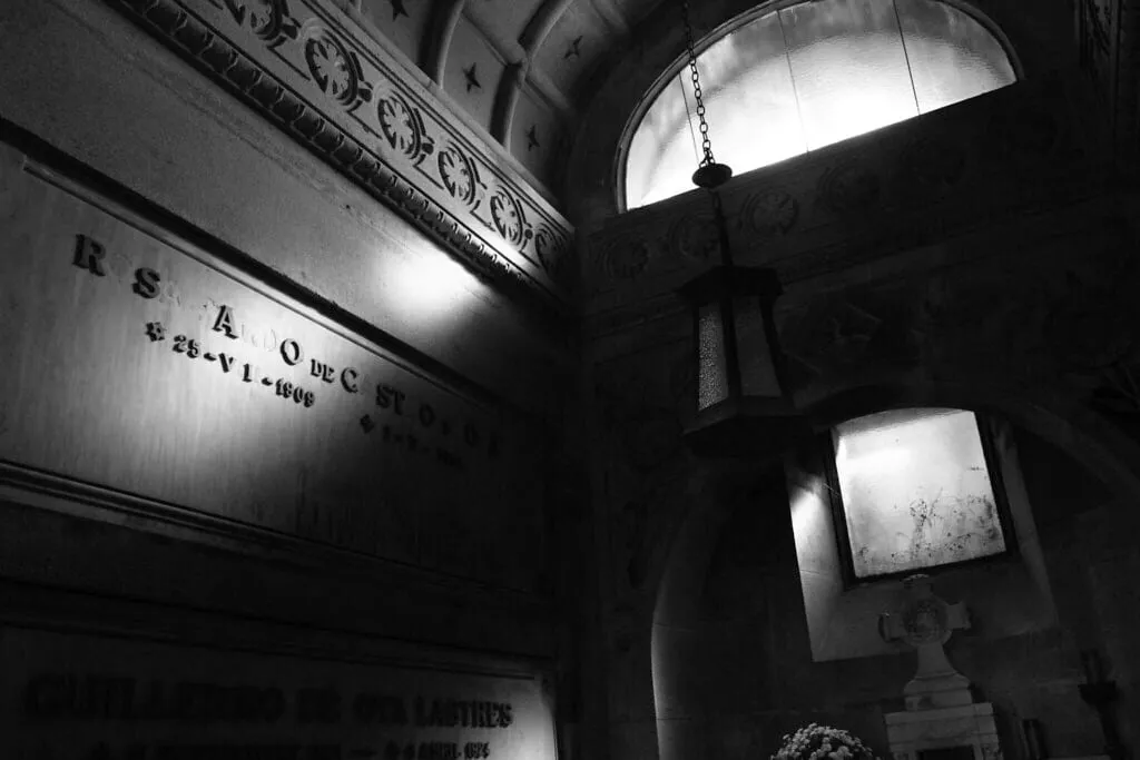 Interior de una cripta con lápidas, una lámpara colgante y un rayo de luz que entra por una ventana alta