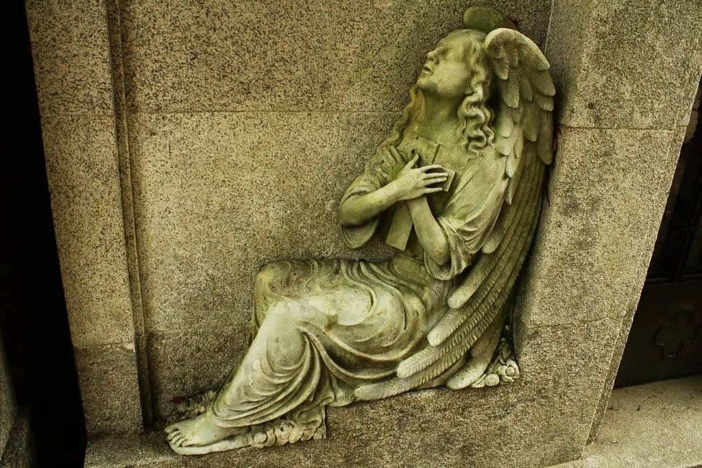 Ángel de piedra recostado en un muro, con un libro entre las manos y la mirada elevada