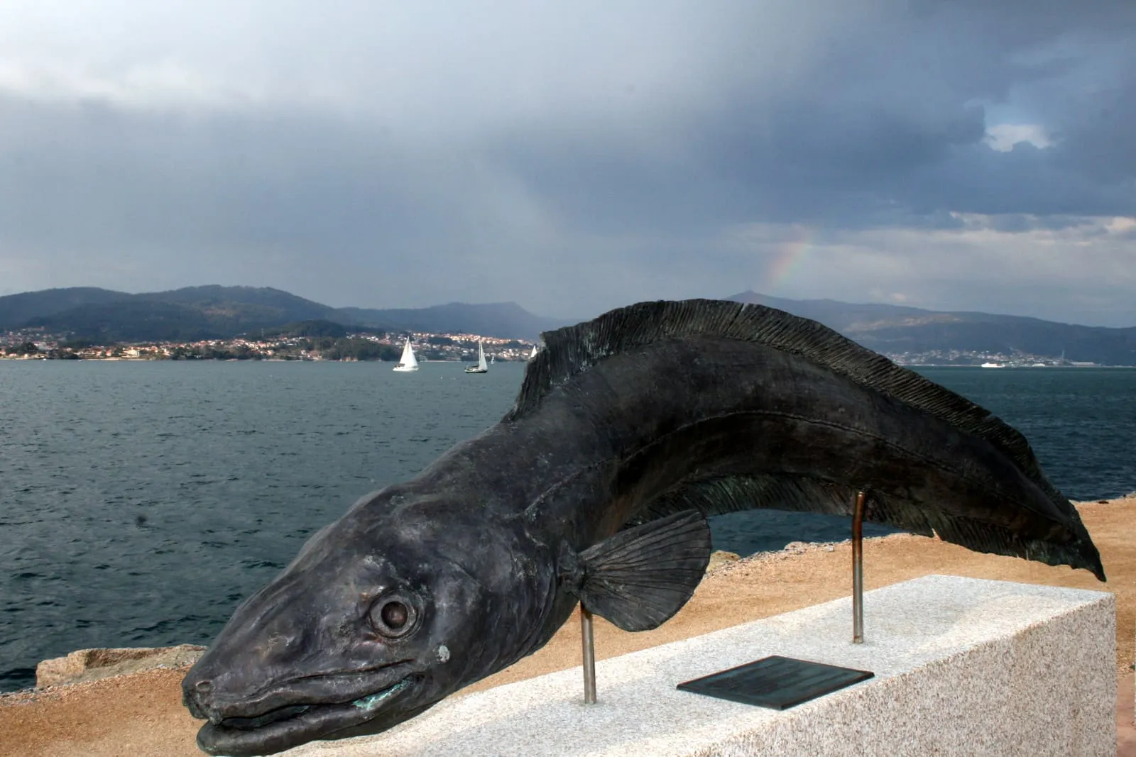 Escultura de bronce de un congrio sobre una peana de granito, con la ría de Vigo al fondo