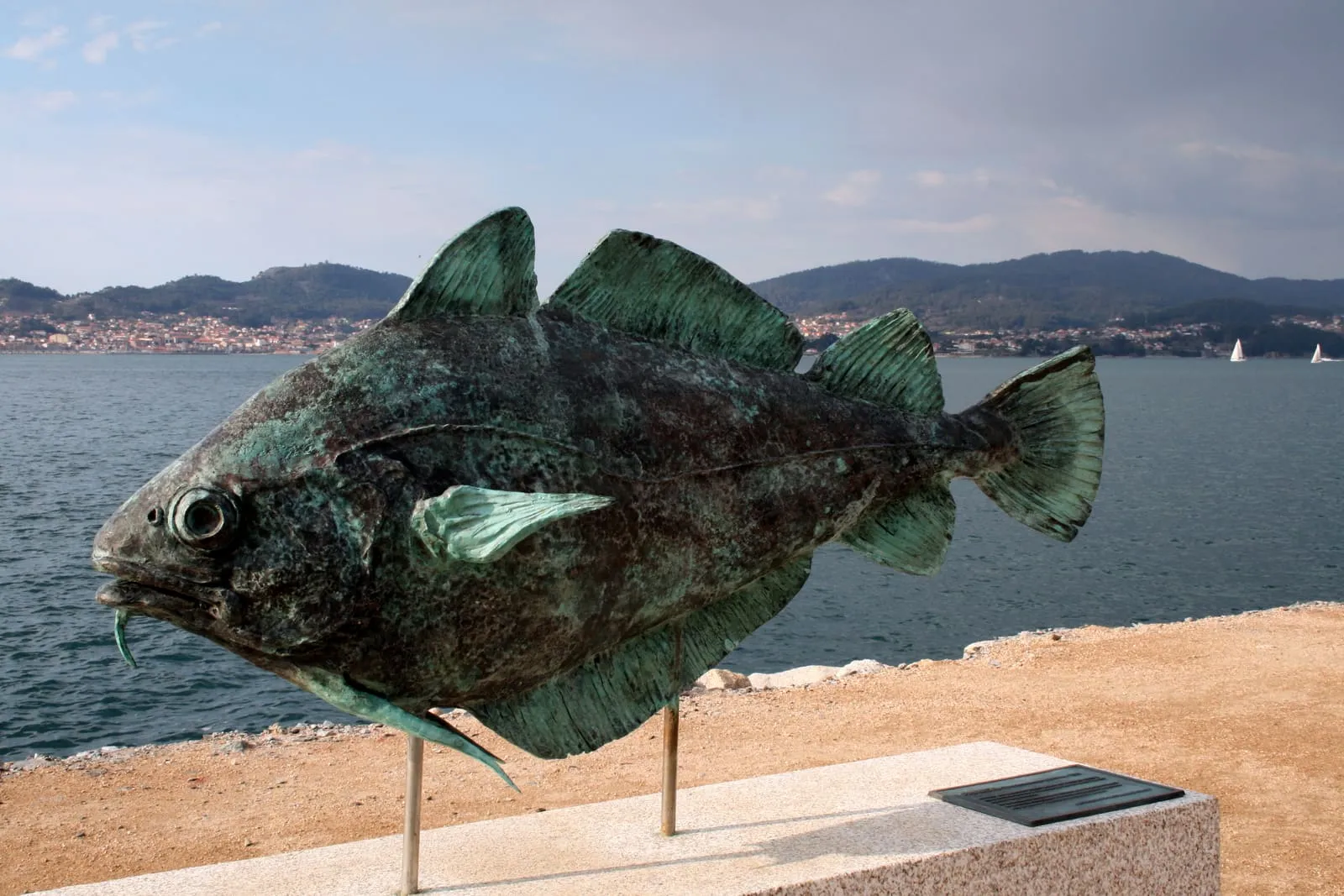 Escultura de bronce de una faneca con pátina verdosa frente a la ría de Vigo